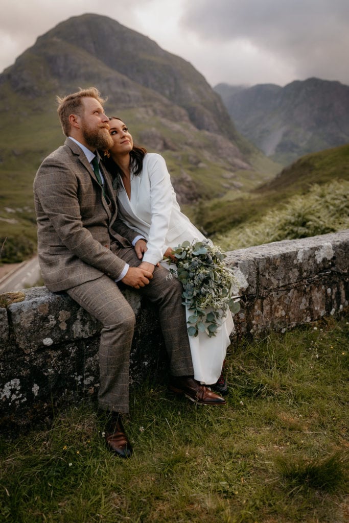 Eloping bride and groom sitting on stone wall in Glencoe valley